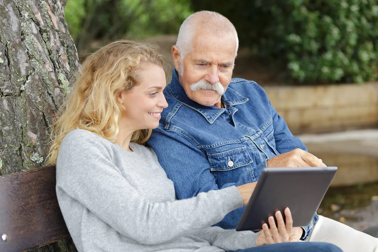 Ein Bild welches einen alten Mann und eine Frau zeigt. Die Frau zeigt dem Mann etwas auf einem Tablet.